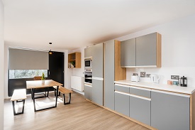 Shared kitchen for townhouses and en suite rooms. Table and benches to the left and kitchen surfaces and oven and microwave to the right. Window in the rear of the photo with a blind. Radiator and shelf and cupboard space also visible.